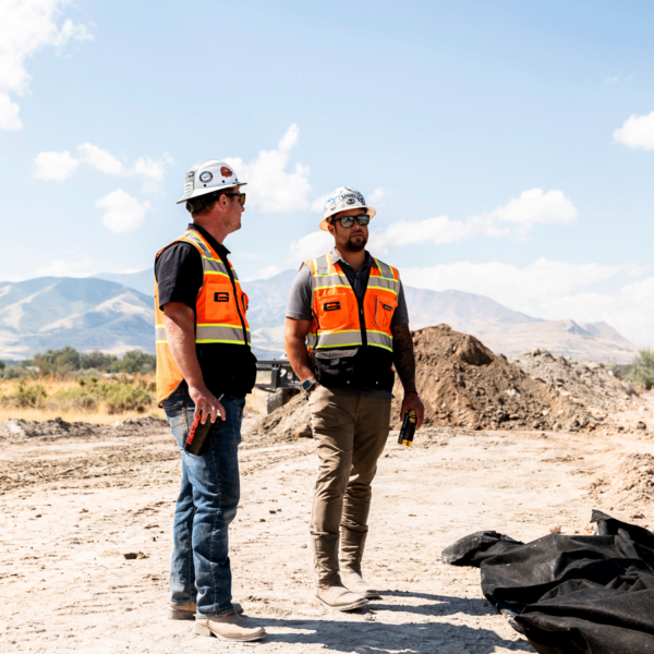 MC Green Employees standing, talking at job site, orange safety vests, white hard hats, mountains in background